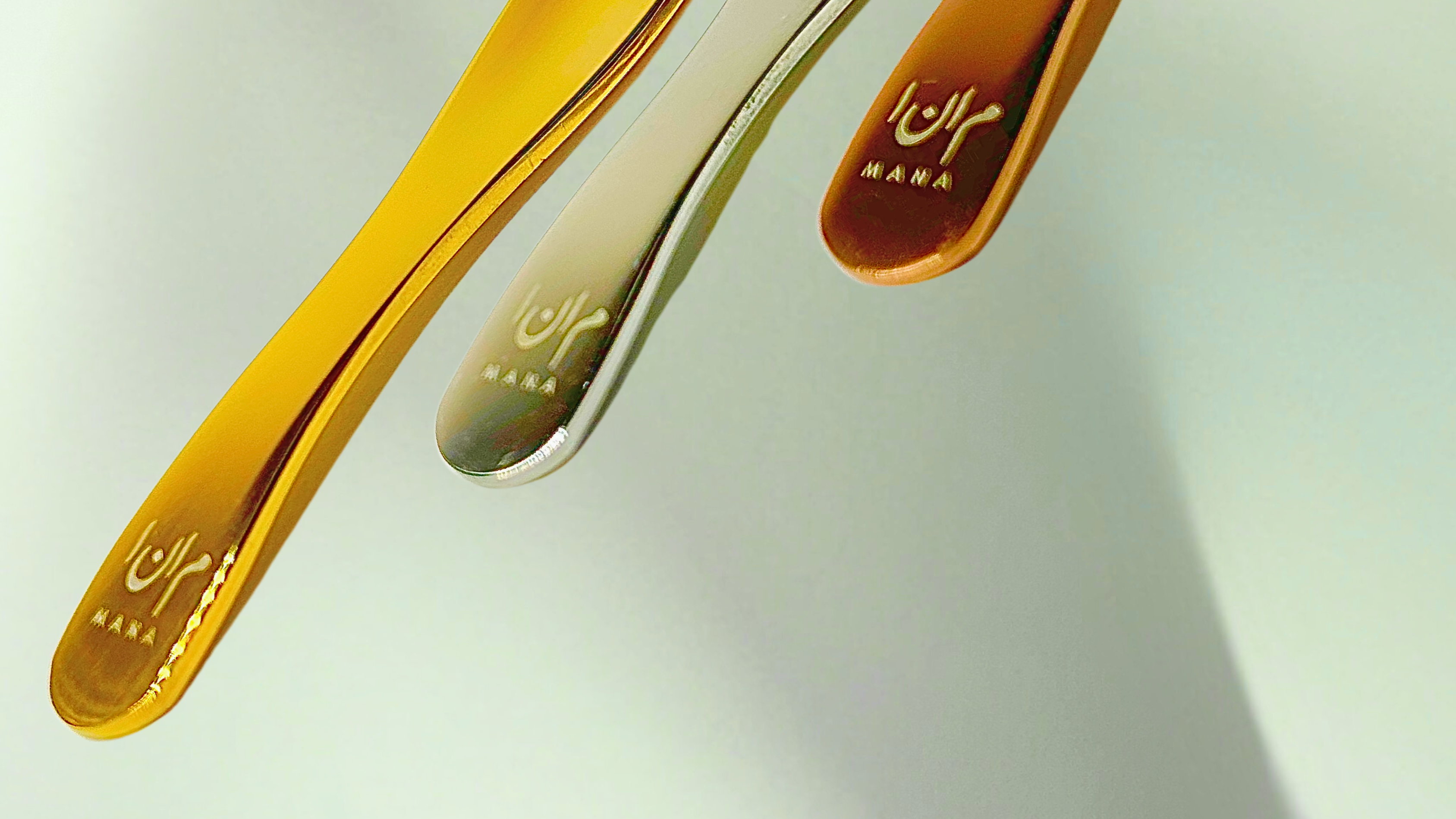 Three colored skincare cosmetic spoon spatulas on a light grayish-green background with the Mana Beauty and Skincare logo engraved into the handles.