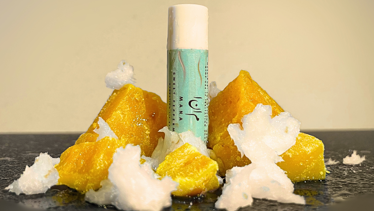 Mana Beauty and Skincare Vitamin E Peppermint Hydrating Lip Balm standing surrounded with beeswax and coconut oil chunks on a dark surface, with a vanilla and water mist sprayed on the landscape and product.